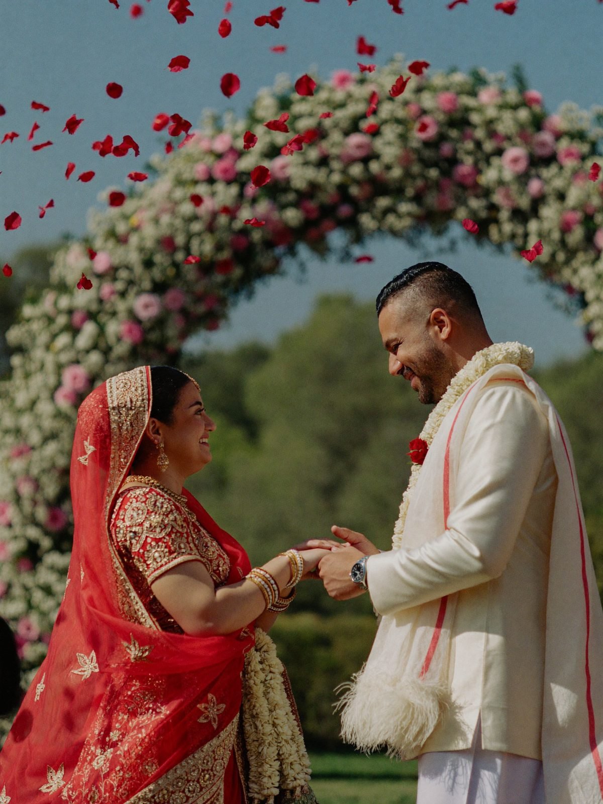 Couple smiling at each other during the varmala as rose petals rain around them, floral arch behind